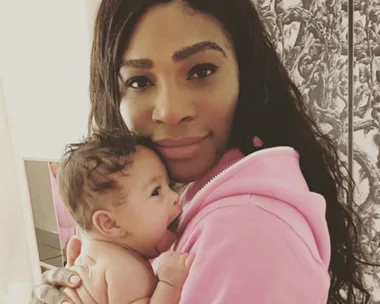 A woman in a pink hoodie holds a newborn baby. Both are looking at the camera.