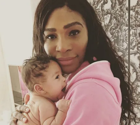 A woman in a pink hoodie holds a newborn baby. Both are looking at the camera.