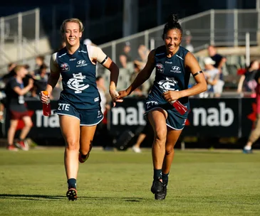 Two female Carlton AFLW players in blue uniforms running and smiling while holding red drink bottles on a sports field.