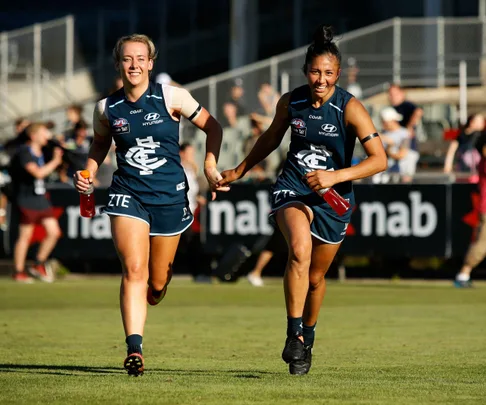 Two female Carlton AFLW players in blue uniforms running and smiling while holding red drink bottles on a sports field.