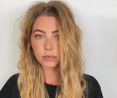 Blonde woman with wavy hair, light blue eyes, and a neutral expression, wearing a black shirt against a plain background.