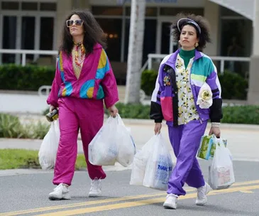 Two women in colorful retro tracksuits walk down a street carrying grocery bags.