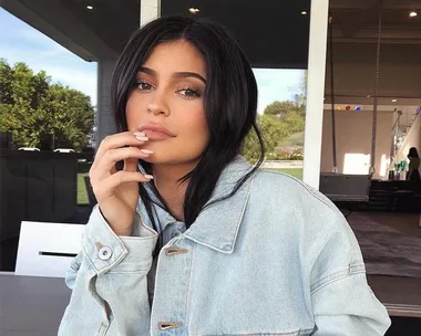 A woman with long black hair, wearing a light denim jacket, sits at a table with her hand near her face, looking at the camera.