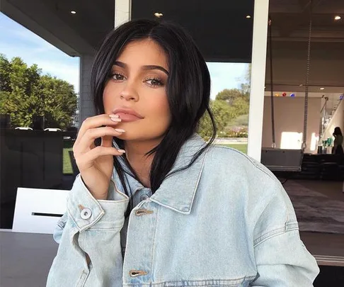 A woman with long black hair, wearing a light denim jacket, sits at a table with her hand near her face, looking at the camera.