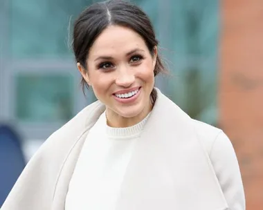 A woman with dark hair tied back, wearing a white top and a light-colored coat, smiling outdoors.