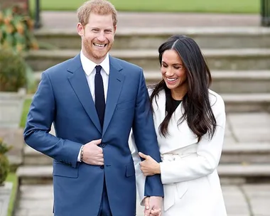 Prince Harry in a blue suit and Meghan Markle in a white coat, smiling and holding hands, on an outdoor stone staircase.