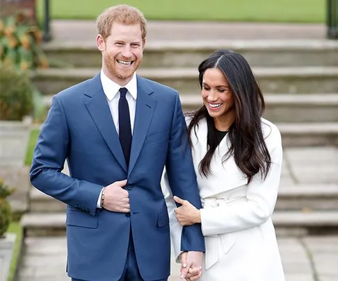 Prince Harry in a blue suit and Meghan Markle in a white coat, smiling and holding hands, on an outdoor stone staircase.