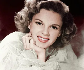 Judy Garland smiling warmly with her chin resting on her hand, wearing a light-colored, translucent blouse.