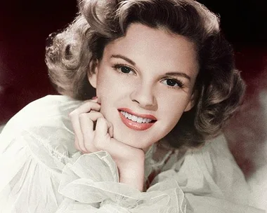 Judy Garland smiling warmly with her chin resting on her hand, wearing a light-colored, translucent blouse.