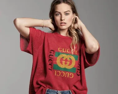 A person in a red Gucci T-shirt and jeans looks at the camera with hands on their head against a plain background.