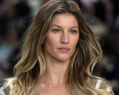 A woman with long, wavy blonde hair, wearing a beige top, appears to be on a runway with a blurred audience in the background.