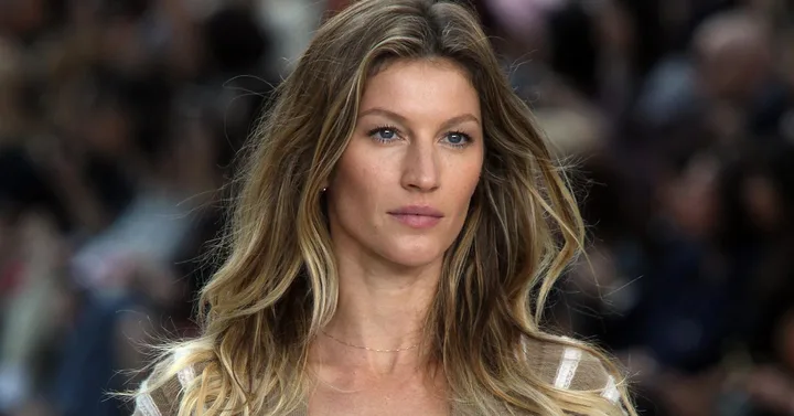 A woman with long, wavy blonde hair, wearing a beige top, appears to be on a runway with a blurred audience in the background.