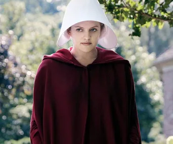 A female character in "The Handmaid's Tale" wearing a white bonnet and red cloak stands outdoors with a serious expression.