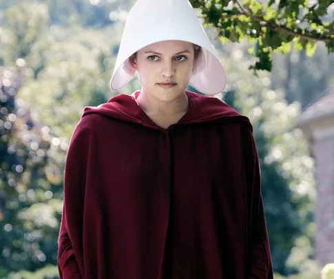 A female character in "The Handmaid's Tale" wearing a white bonnet and red cloak stands outdoors with a serious expression.