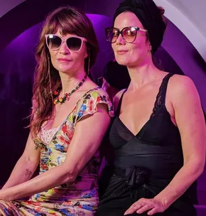 Two women wearing sunglasses, the left in a colorful dress and the right in a black outfit with a headwrap, sit in front of a purple background.