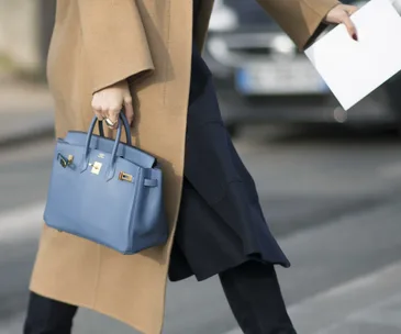 Person in a camel coat carrying a blue handbag and holding a white paper, walking on a street.