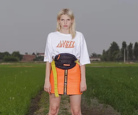 Blonde woman in a white "Angel" shirt and orange skirt, standing in a field, wearing a black belt bag with a yellow strap.