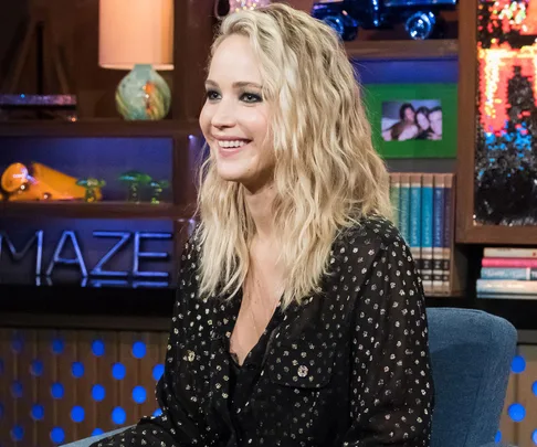 Jennifer Lawrence smiling in a black outfit with a sparkling design, seated in a talk show set with books and decorations in the background.