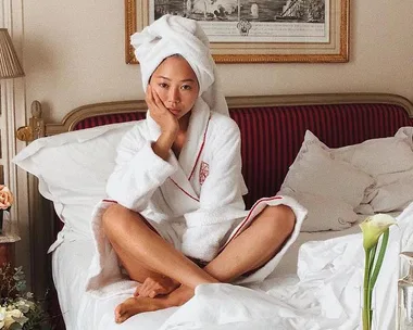 A woman in a bathrobe and towel on her head sits on a bed in a luxurious room, surrounded by skincare products.