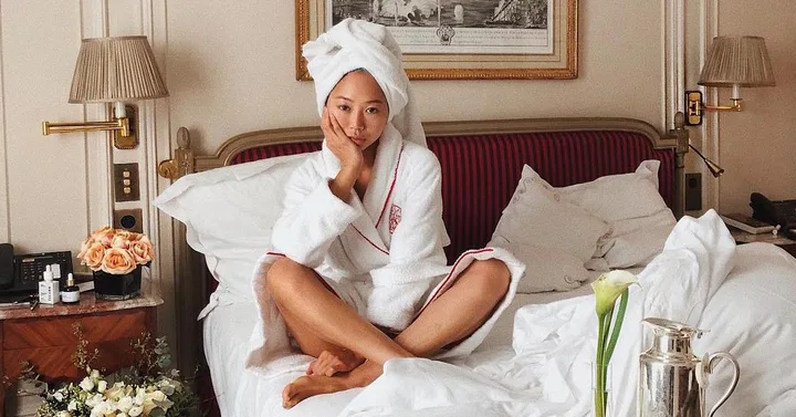 A woman in a bathrobe and towel on her head sits on a bed in a luxurious room, surrounded by skincare products.