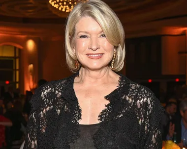 Martha Stewart in a black lace outfit, smiling at a formal event with warm lighting and a chandelier in the background.