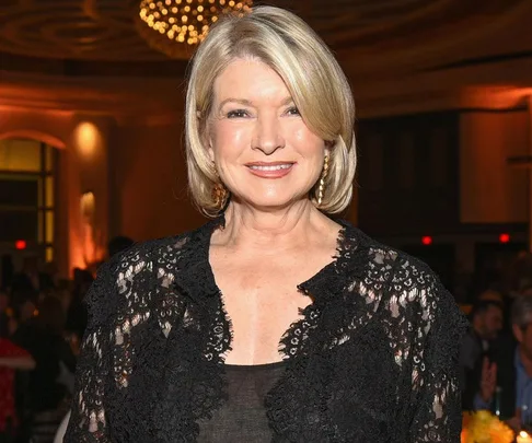 Martha Stewart in a black lace outfit, smiling at a formal event with warm lighting and a chandelier in the background.