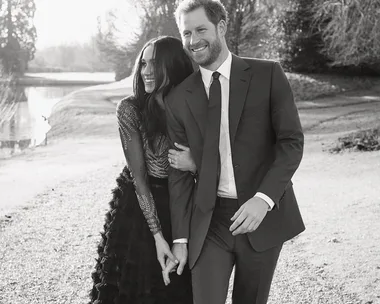 Meghan Markle and Prince Harry walking arm in arm, smiling, in a black and white photo near a pond and trees.