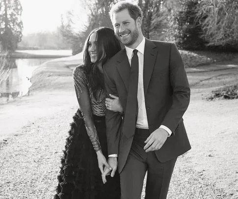 Meghan Markle and Prince Harry walking arm in arm, smiling, in a black and white photo near a pond and trees.