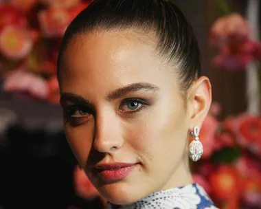 Woman with elegant makeup, earrings, and a sleek hairstyle, posing against a blurred floral background.