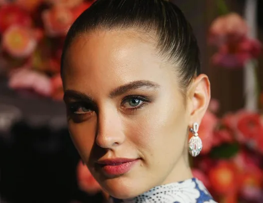 Woman with elegant makeup, earrings, and a sleek hairstyle, posing against a blurred floral background.