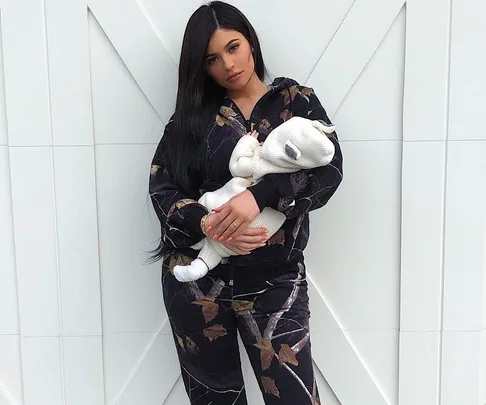 Woman in black floral tracksuit holding a baby dressed in a white outfit, standing against a white geometric background.