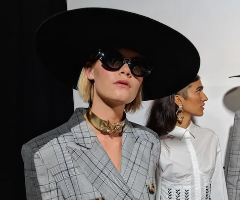 Two models posing; one in a wide-brimmed hat, sunglasses, gray checkered blazer, gold necklace; the other in a white shirt with earrings.