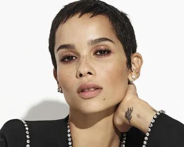 Close-up of Zoë Kravitz with short hair, multiple earrings, and a tattoo on her hand, posing with her hand resting on her neck.