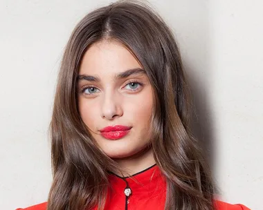 A woman with long brown hair and bright red lipstick wearing a red top against a neutral background.