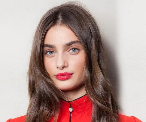 A woman with long brown hair and bright red lipstick wearing a red top against a neutral background.