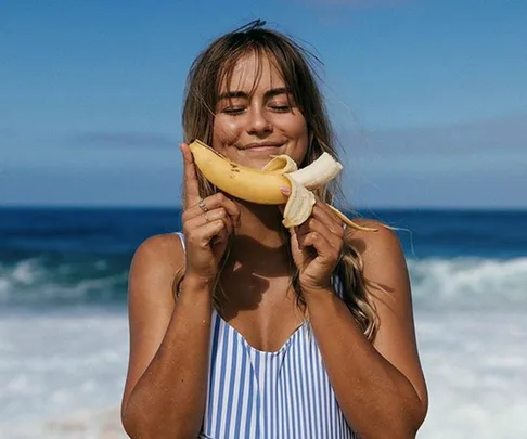 Woman in striped swimsuit at beach holding peeled banana, smiling with closed eyes against a backdrop of ocean waves and blue sky.