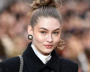 A model on the runway with hair in a high bun wearing bold statement earrings and a black coat.