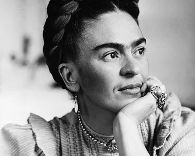 Black and white portrait of Frida Kahlo, with braided hair and resting head on hand, showcasing her iconic unibrow and attire.