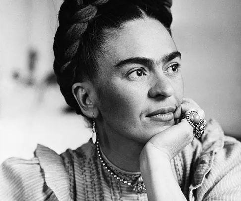 Black and white portrait of Frida Kahlo, with braided hair and resting head on hand, showcasing her iconic unibrow and attire.