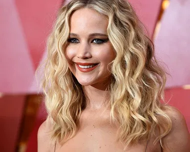 Blonde actress with wavy hair, dramatic eyeliner, and a red dress on the 2018 Oscars red carpet against a pink background.