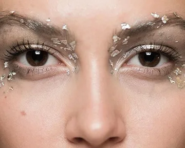 Close-up of a person's eyes with metallic flakes applied as makeup around the eyebrows and eyelids.