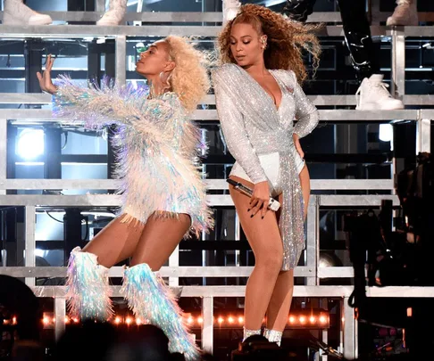 Two performers in sparkling outfits dancing on stage at Coachella; one wearing a bodysuit with a cape, the other in fringed attire.