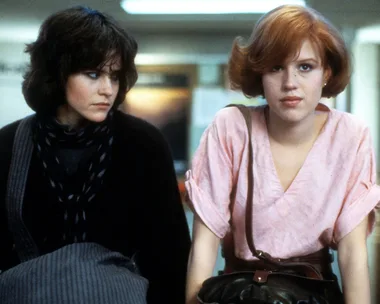 Two teenage girls from "The Breakfast Club" sitting together; one wears black, and the other is in pink with red hair.