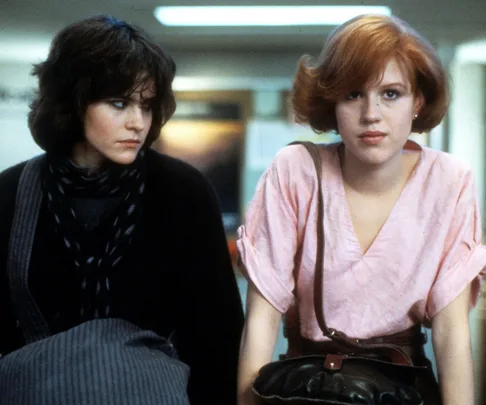 Two teenage girls from "The Breakfast Club" sitting together; one wears black, and the other is in pink with red hair.