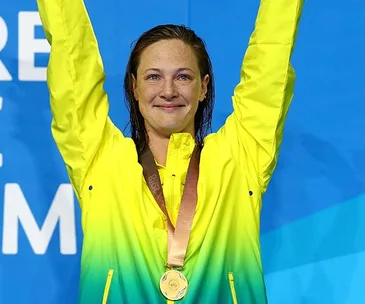 Woman in a yellow-green jacket holding her arms up with a medal around her neck, standing in front of a blue background.