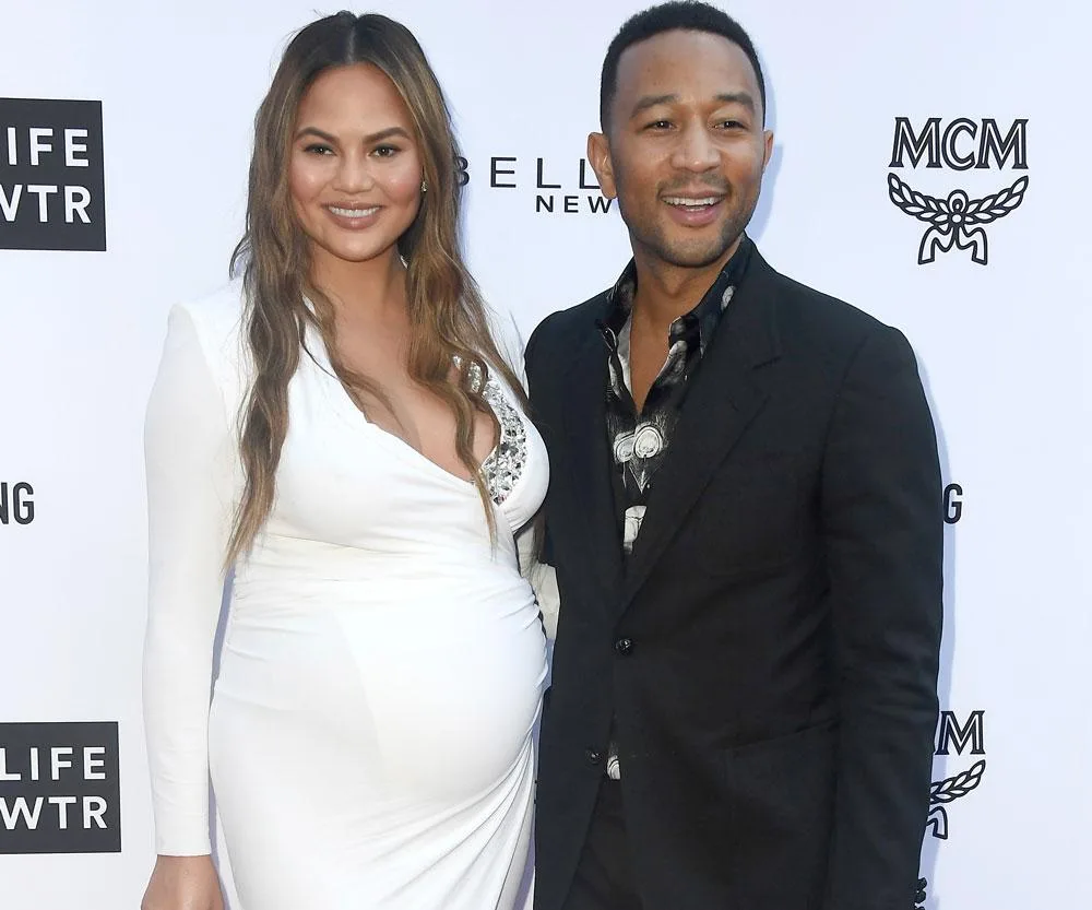 Chrissy Teigen and her husband John Legend smiling together at her baby shower, with Chrissy in a white dress.

