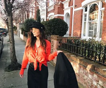 A woman in a red blouse and black skirt walks on a tree-lined street, holding a black jacket in one hand.