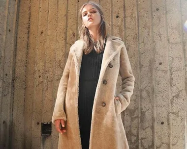 Woman wearing a long beige teddy coat stands against a concrete wall.