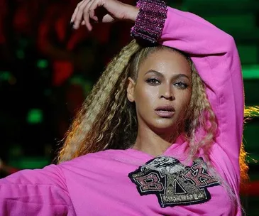 Beyoncé performing at Coachella in a pink sweatshirt with a Greek-letter patch and sequined cuffs, her hand raised above her head.