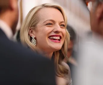 Elisabeth Moss smiling at a public event, wearing pearl earrings and red lipstick.